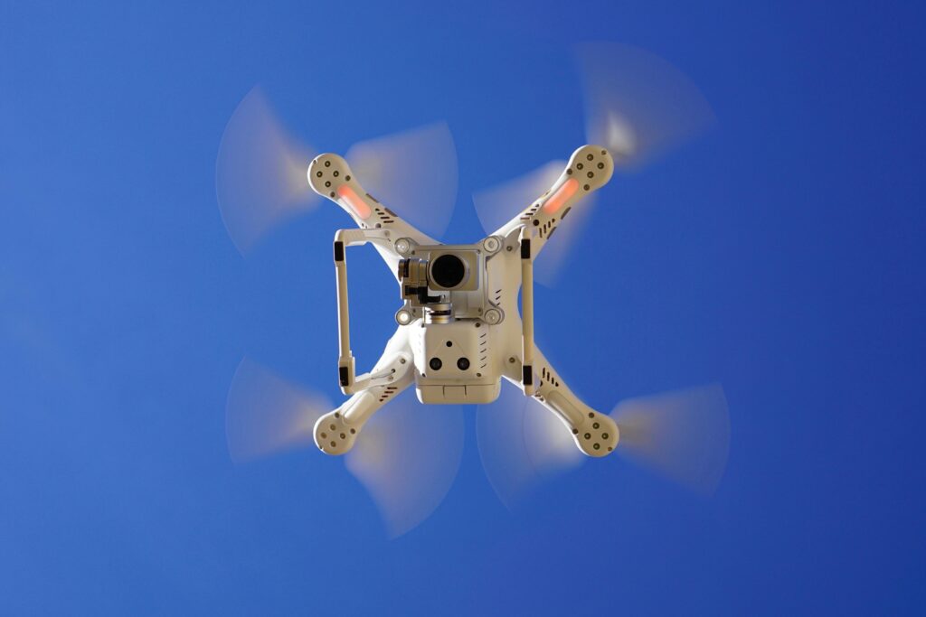 Aerial view of white drone flying with rotating blades against a clear blue sky, capturing video footage.