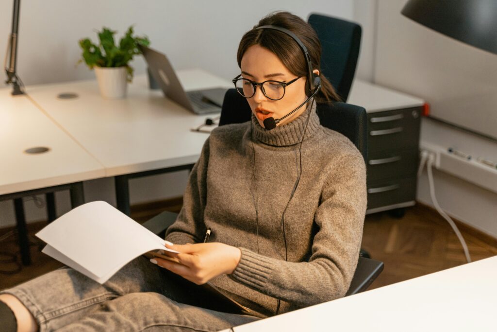 Professional woman in an office wearing a headset and reading documents at a call center desk.